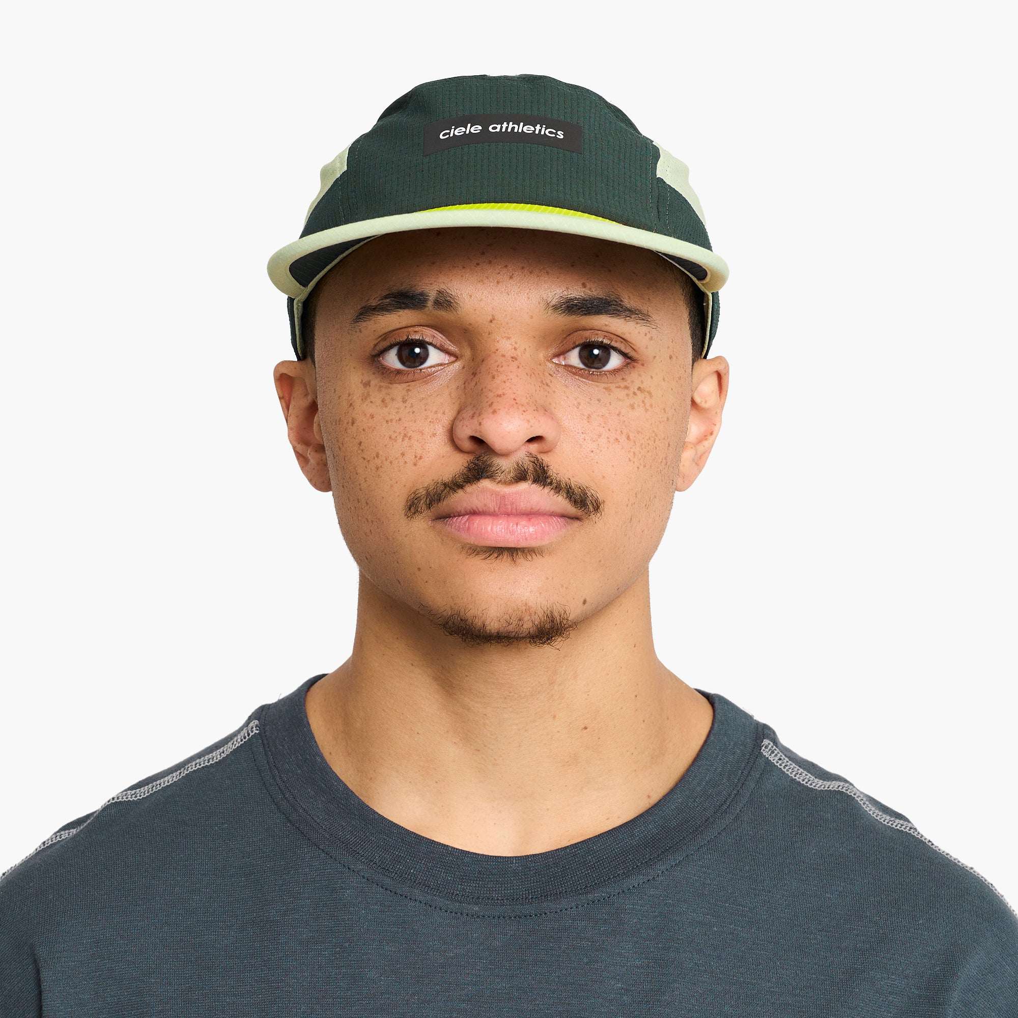 Ciele Field running cap on model: dark green body, cream front patch, neon yellow brim edge